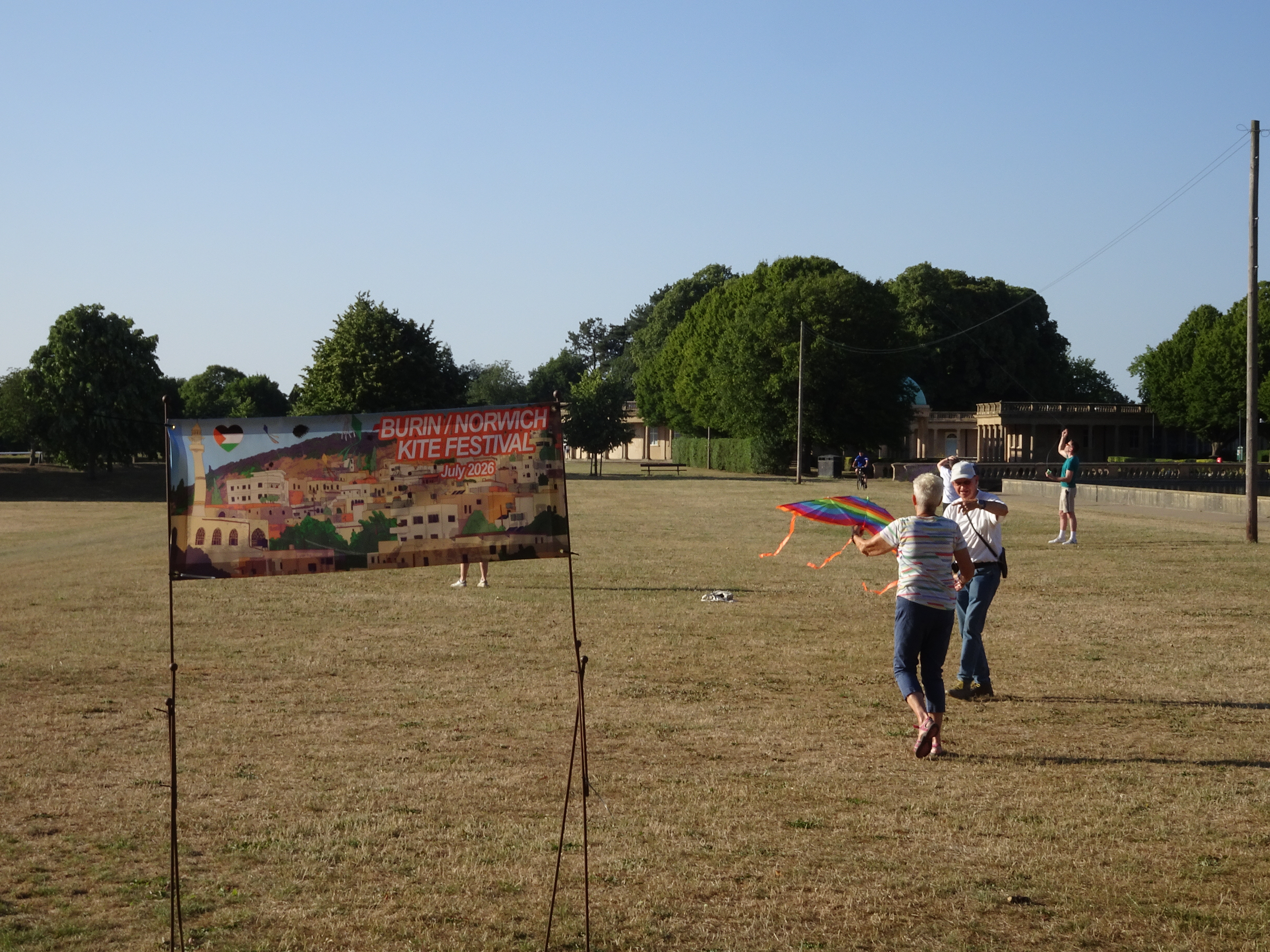 A view of Eaton Park with trees and the central park buildings in the background, a banner for the festival in the foreground and two people launching a kit just to the right.