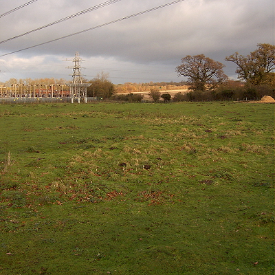 The site of Arminghall Henge, with electricity substation in the background, wires overhead, and a an arc of rougher vegetation marking part of the inner ditch amongst the grass.