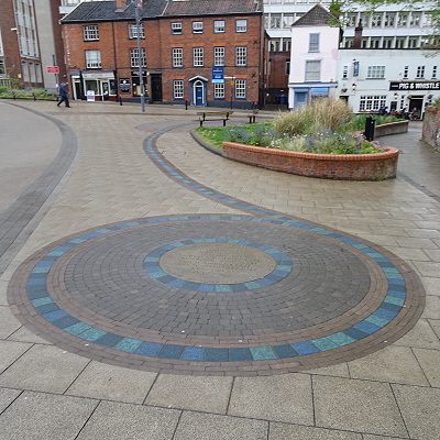 The pedestrianised and paved Westlegate, looking towards All Saints Green, with the Pig and Whistle pub visible in the background behind the seats and flower beds, and in the foreground a circular pattern with grey and varied blue setts, which then head off in a sinuous line down Westlegate.
