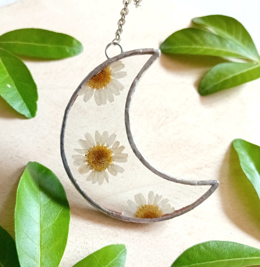 A crescent-shaped clear glass pendant encapsualting daisy flowers, with leaves in the background of the picture. Picture by Gemma and Mychael Pash.