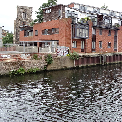 Where the Dalymond once entered the Wensum, at the end of Hansard Lane, off Fishergate: with a concrete block between a wall and pilings, modern flats behind to the right, a small brick building covered in graffiti behind, a railed car park to the left, and the square flinten tower of St. Edmund's church in the background.
