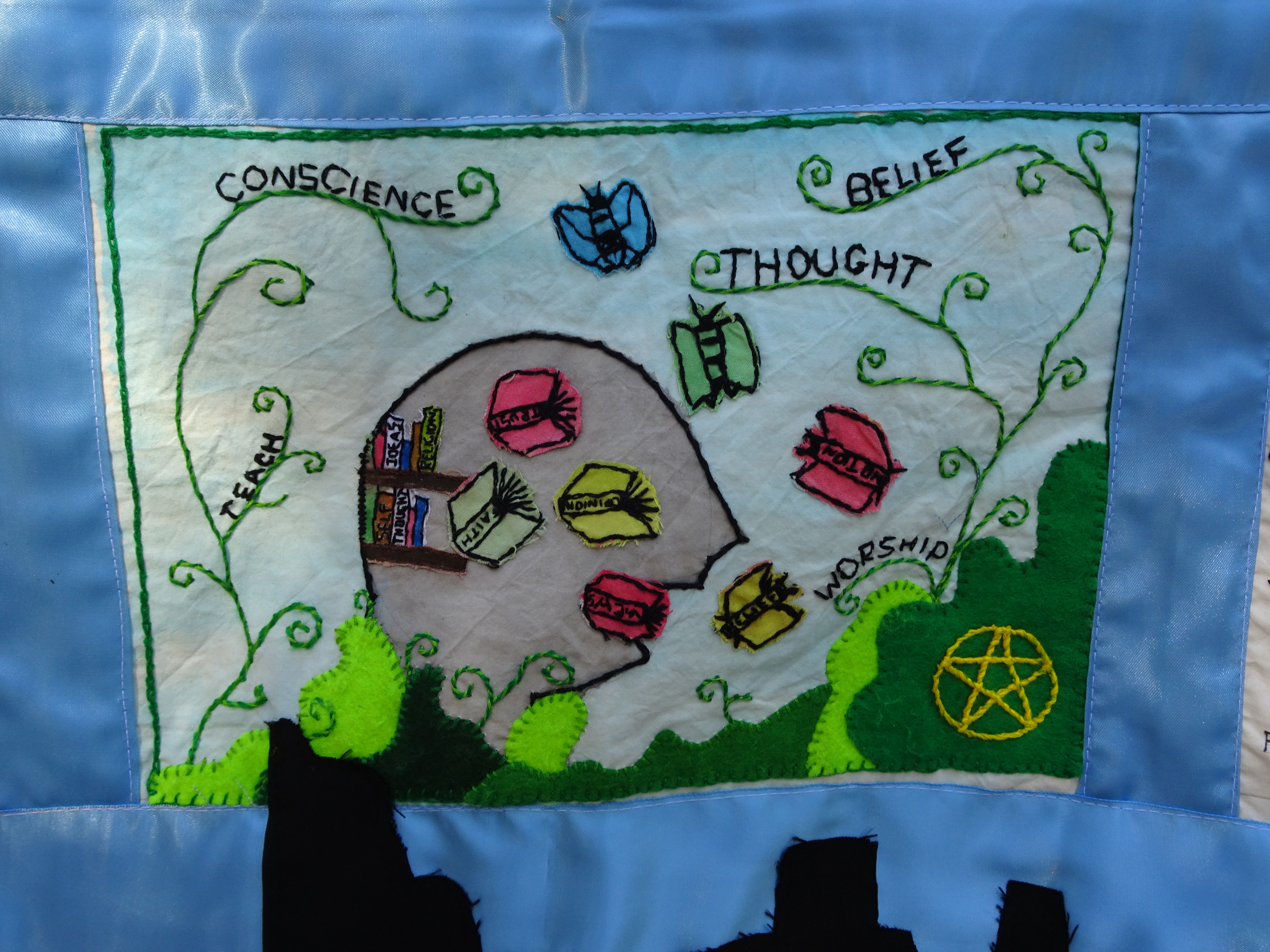 Quilt panel depicting Article 9 of the European Convention on Human Rights, with a picture of a head and book-like butterflies, with the words, 'teach, 'belief', 'thought' and 'worship'.