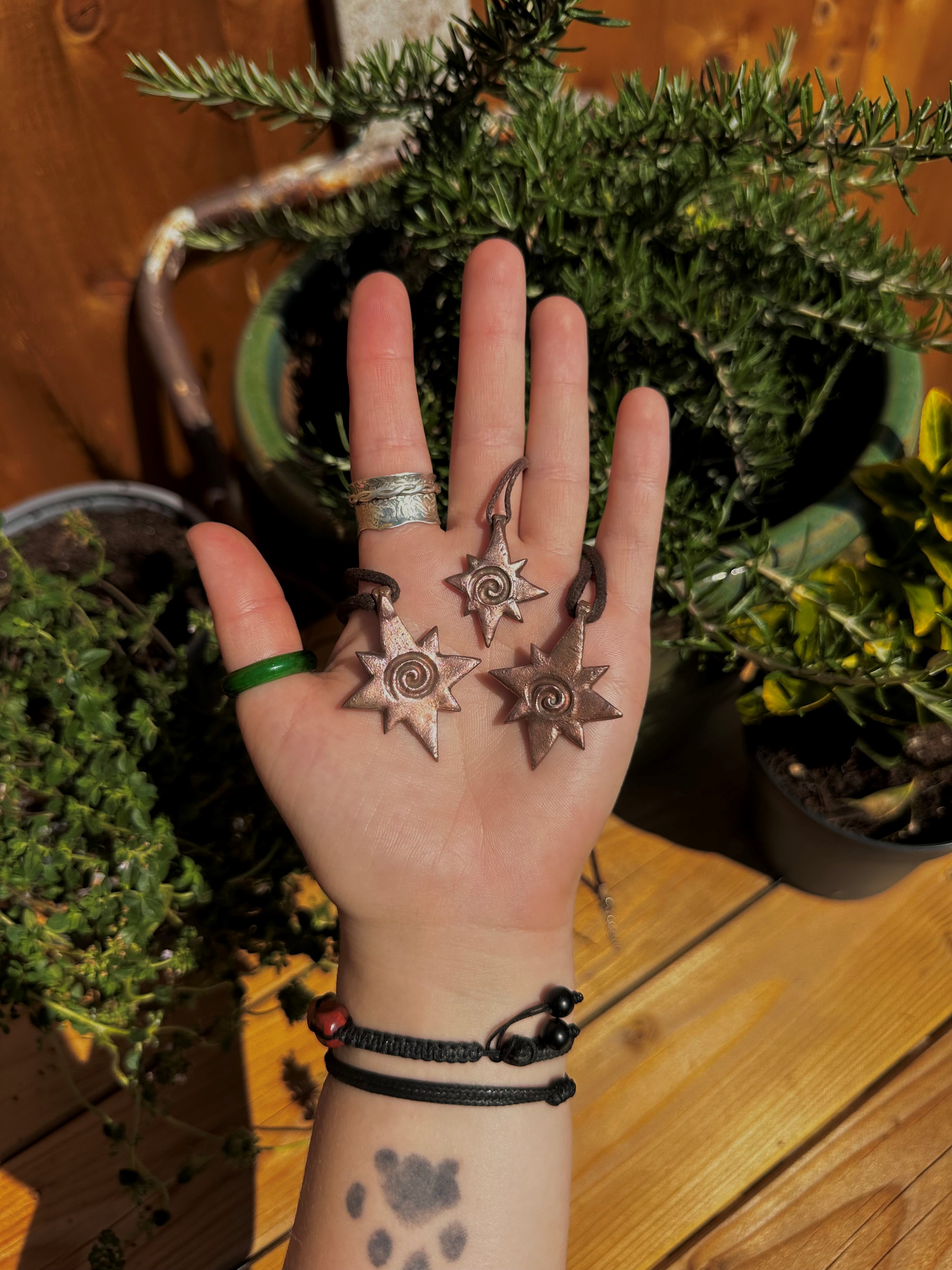 A left hand, with rings, bracelets and a dog paw tattoo on the wrist, holding three pendants in star shape with spiral centres, in front of a pot of rosemary. Picture by Jodie Hyson.