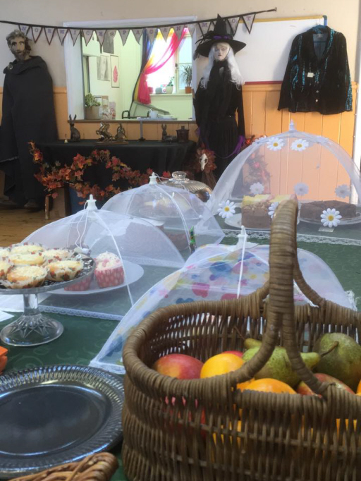 A table with cakes and a basket of apples and pears, with the room beyond and the mannekins of Flint and Hazel in the background by the tea hatch, with a table ofBoudica-related items and bunting spelling out 'Norwich Pagan Moot' between them.