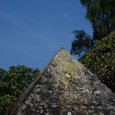 The top of a pyramidal grave marker pointing at the moon, with a tree behind.