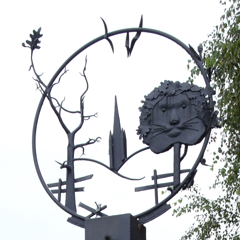 The Thorpe Hamlet village sign in Norwich: a ring with the cathedral, broken fences, trees, birds and a 'foliate lion head'.