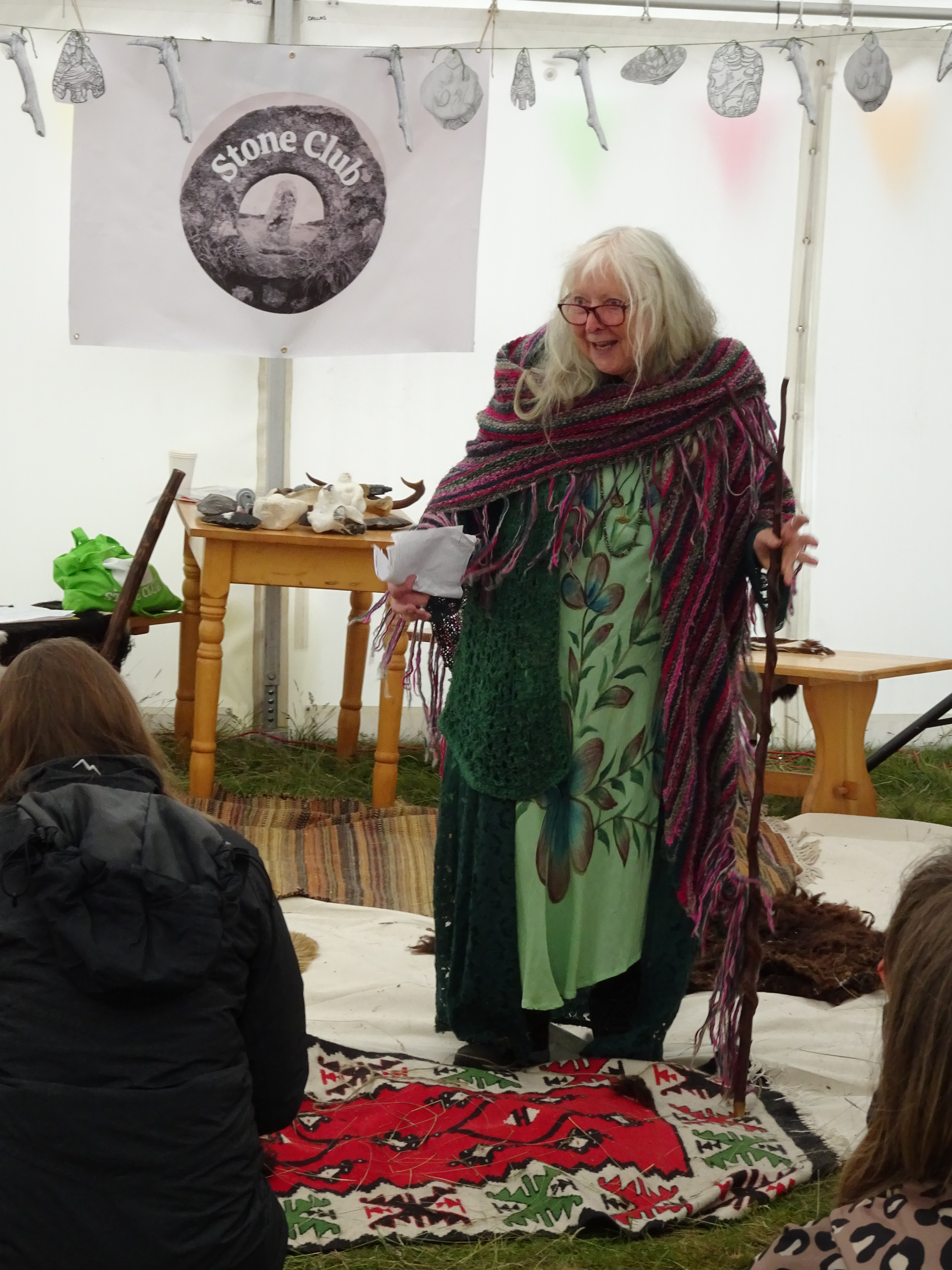 Val Thomas speaking at the Grimes Graves Flint Festival, 'Beneath the Surface', on 15th June 2024, standing with her forked stick in front of the audience in the talks tent, with the 'Stone Club' banner and a table of replica neolthic artefacts behind her.